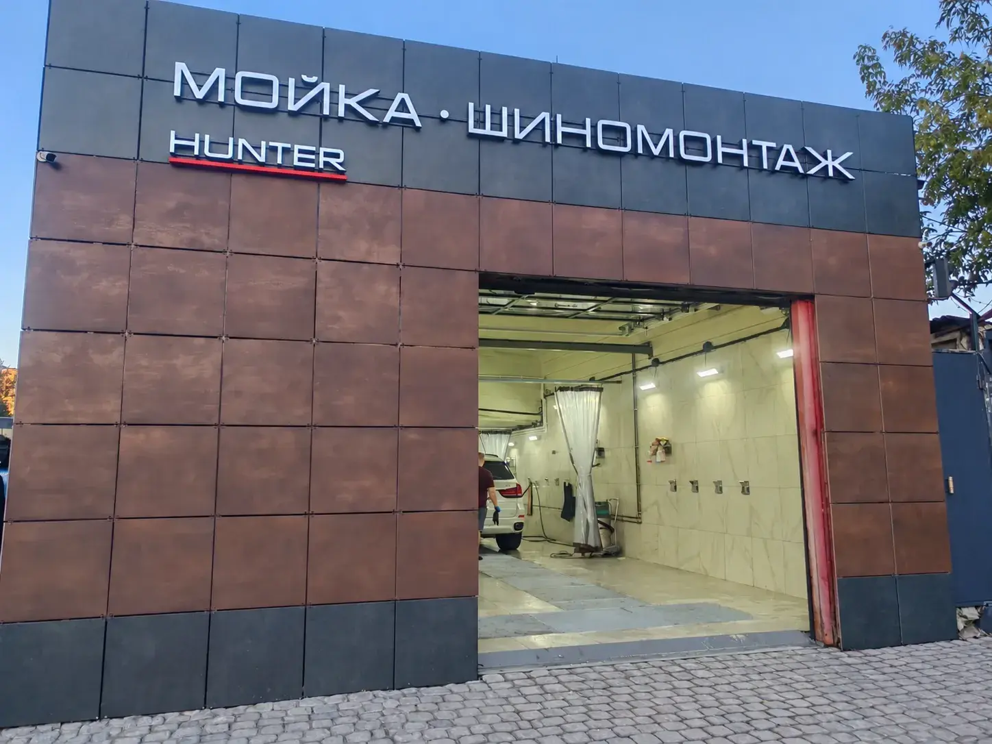 Фасад в плитке под медь с вывеской «Мойка · Шиномонтаж» и надписью Hunter, открытый въезд в бокс