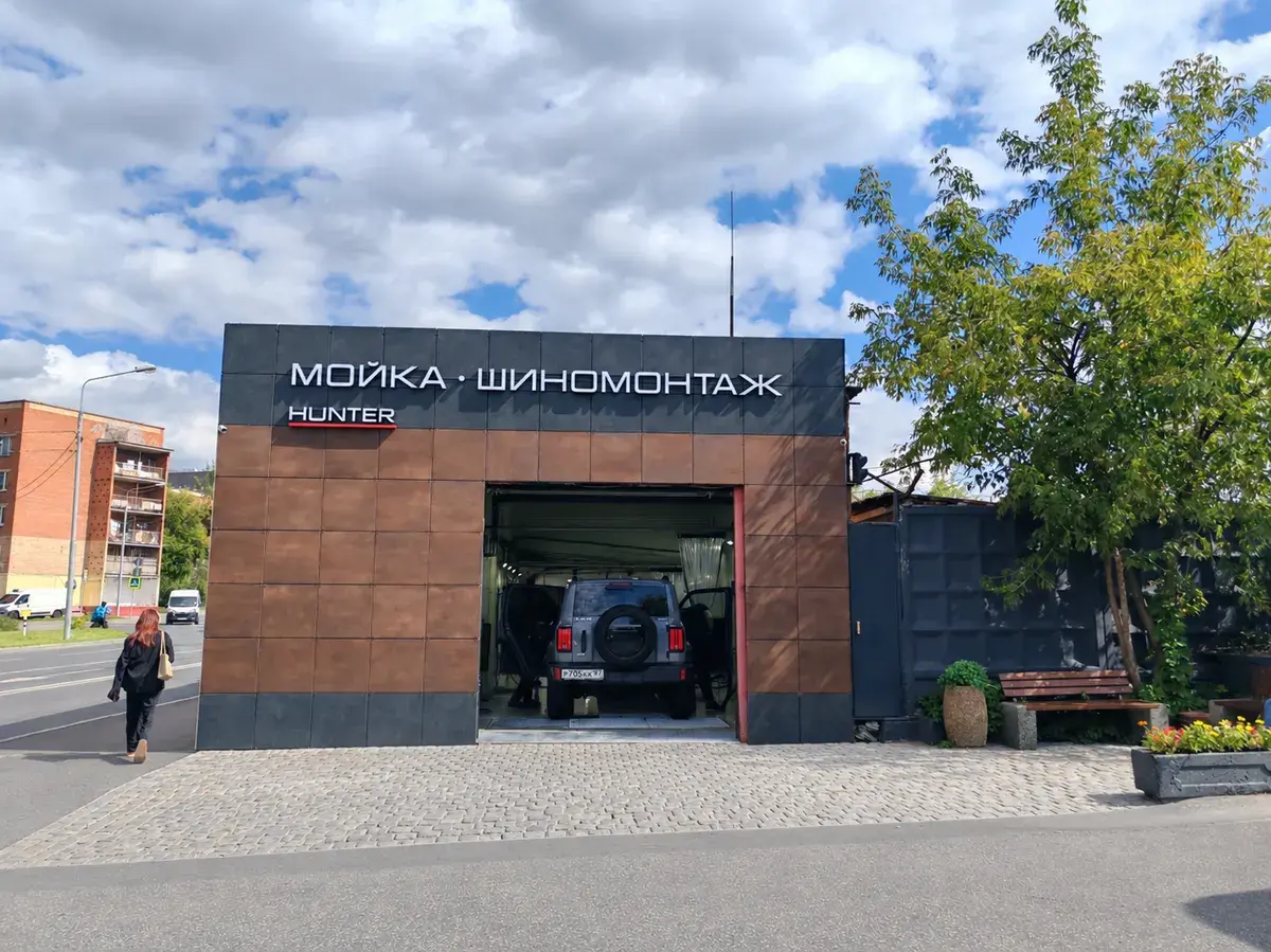Здание «Мойка · Шиномонтаж» днём: облака, дерево, скамейка и клумба у входа