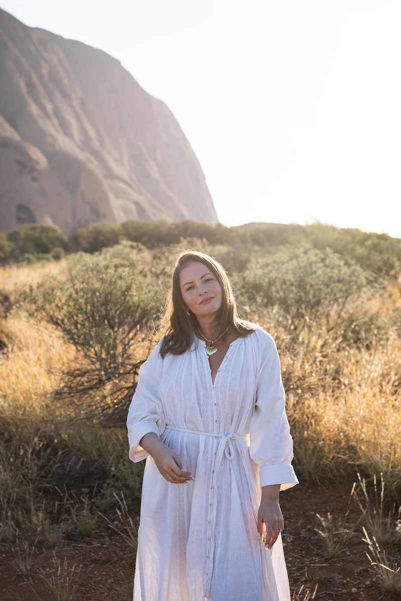 Fernanda stands peacefully in the sunlight with Uluru and scrubland in the background.